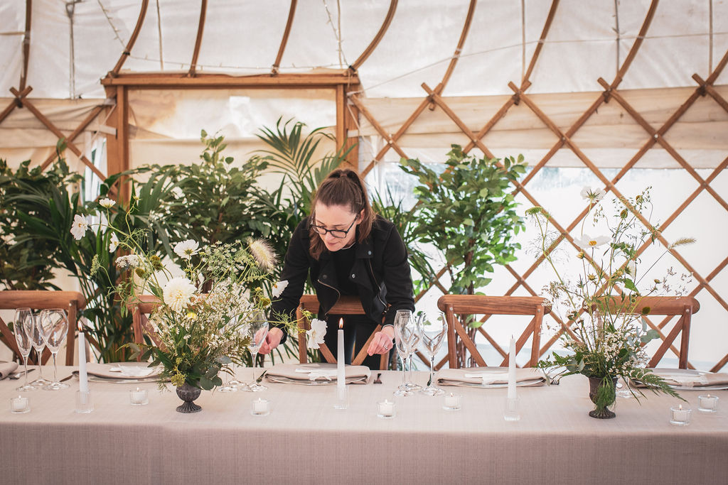 Yurt wedding styled shoot with lots of natural details including leafy green plants, wooden furniture and loose floral arrangements. Hannah a white female wears her long brown hair tied back in a pony tail. Hannah wears large glasses and all black while add the finishing touches to the wedding table.