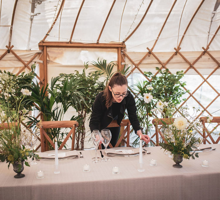 Yurt wedding styled shoot with lots of natural details including leafy green plants, wooden furniture and loose floral arrangements. Hannah a white female wears her long brown hair tied back in a pony tail. Hannah wears large glasses and all black while add the finishing touches to the wedding table.