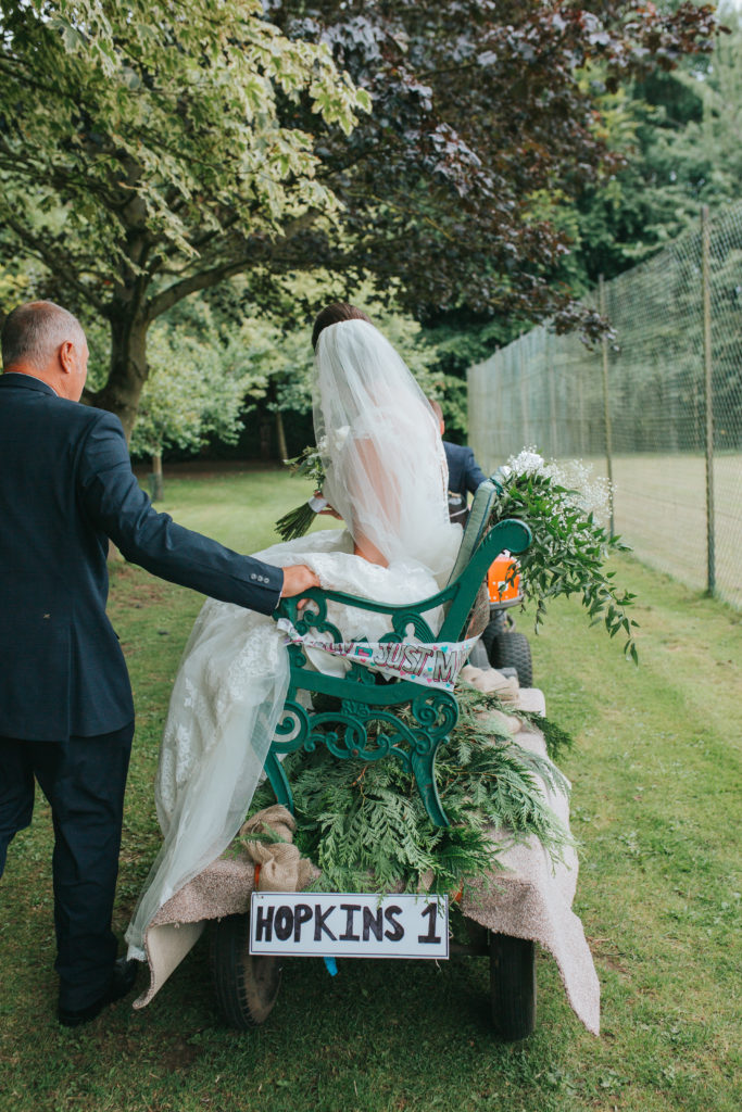 Bride Lucy sits on a bench being towed away by her Groom Sam on a lawn mover.