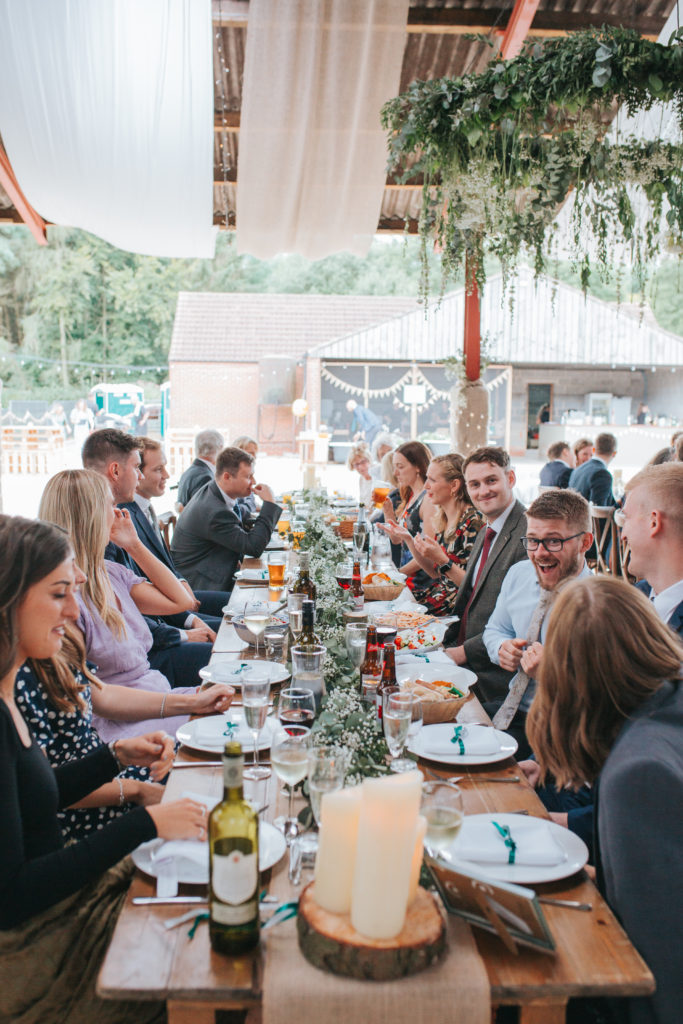 Guests tuck into sharing platters as they dine in the cow shed on long wooden trestle tables. Large floral chandeliers hang above the table.