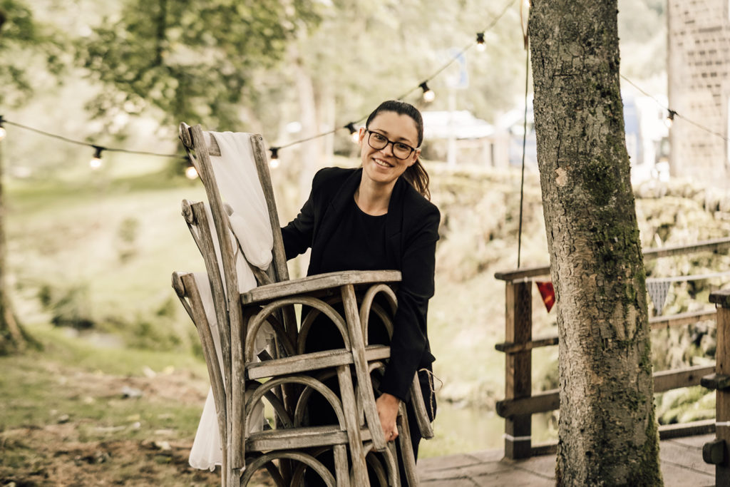 Hannah from Hannah Rachael Weddings smiles as she carries a stack of wooden cross back chairs. Hannah is a white female. She wears her long brown hair tied back in a ponytail, has think black glasses and wears all black. 