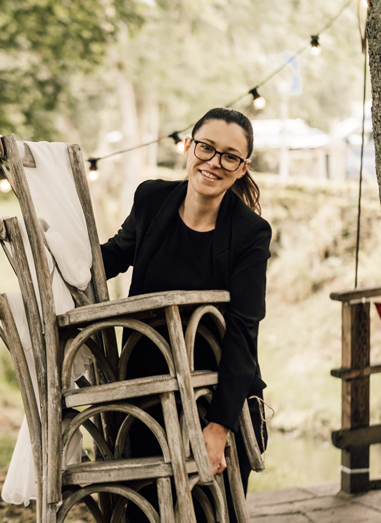 Hannah from Hannah Rachael Weddings smiles as she carries a stack of wooden cross back chairs. Hannah is a white female. She wears her long brown hair tied back in a ponytail, has think black glasses and wears all black.