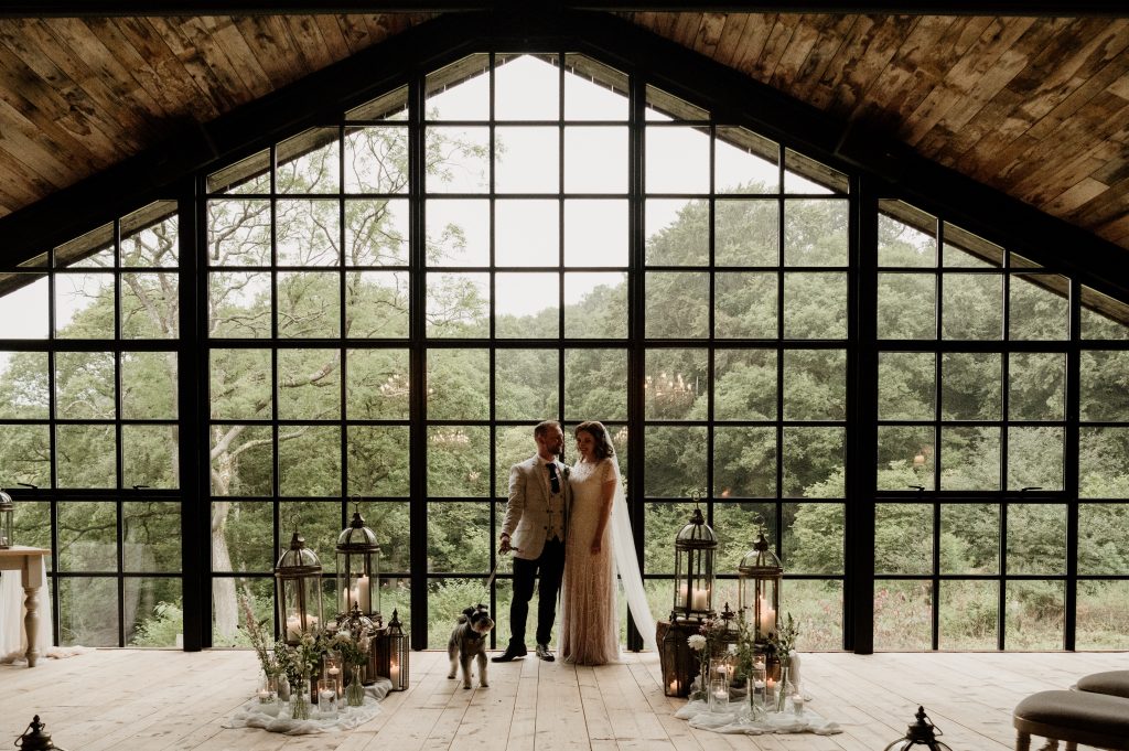 Heather & Phil stand with dog Eddie against a large window backdrop which makes up the mezzanine ceremony space. They are surrounded by lanterns, vases of seasonal blooms and floating candles