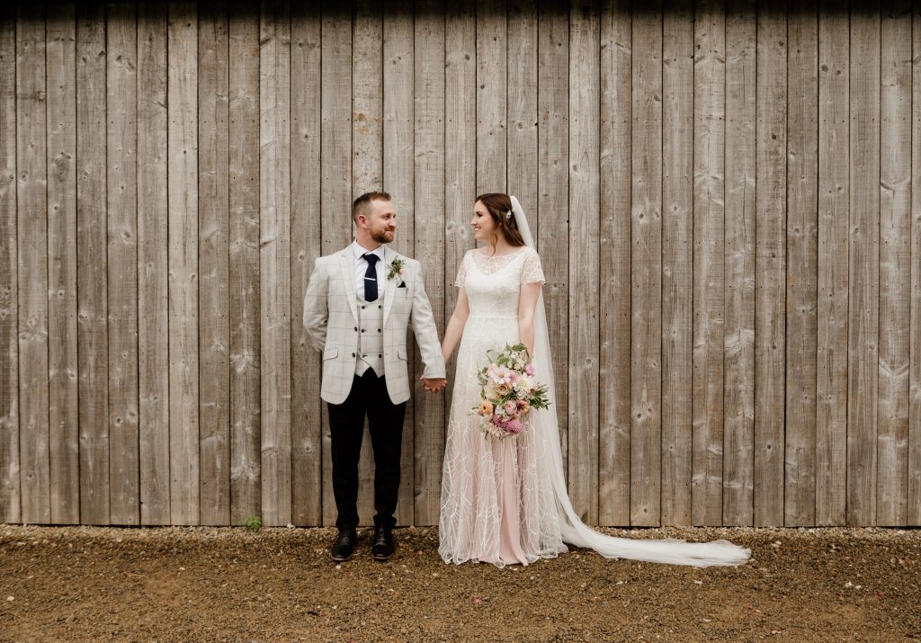 Heather and Phil stand in front of a wooden wall while holding hands.