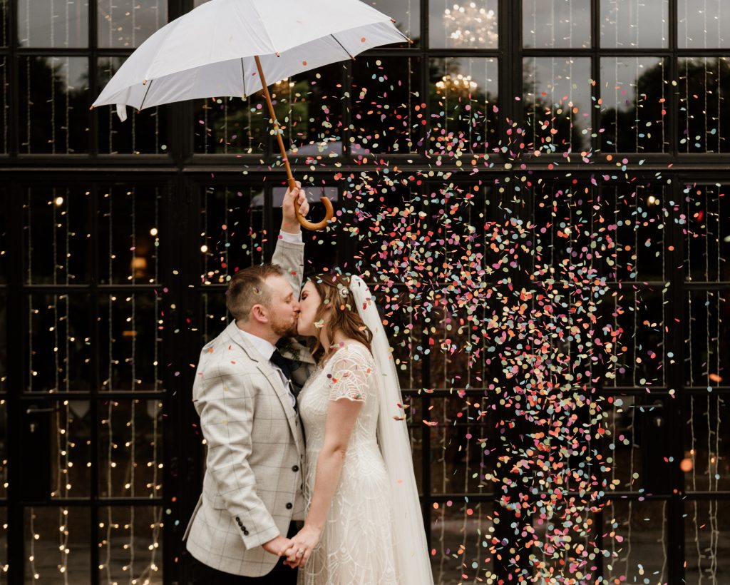 Heather and phil kiss as bright confetti falls around them from under an umbrella.
