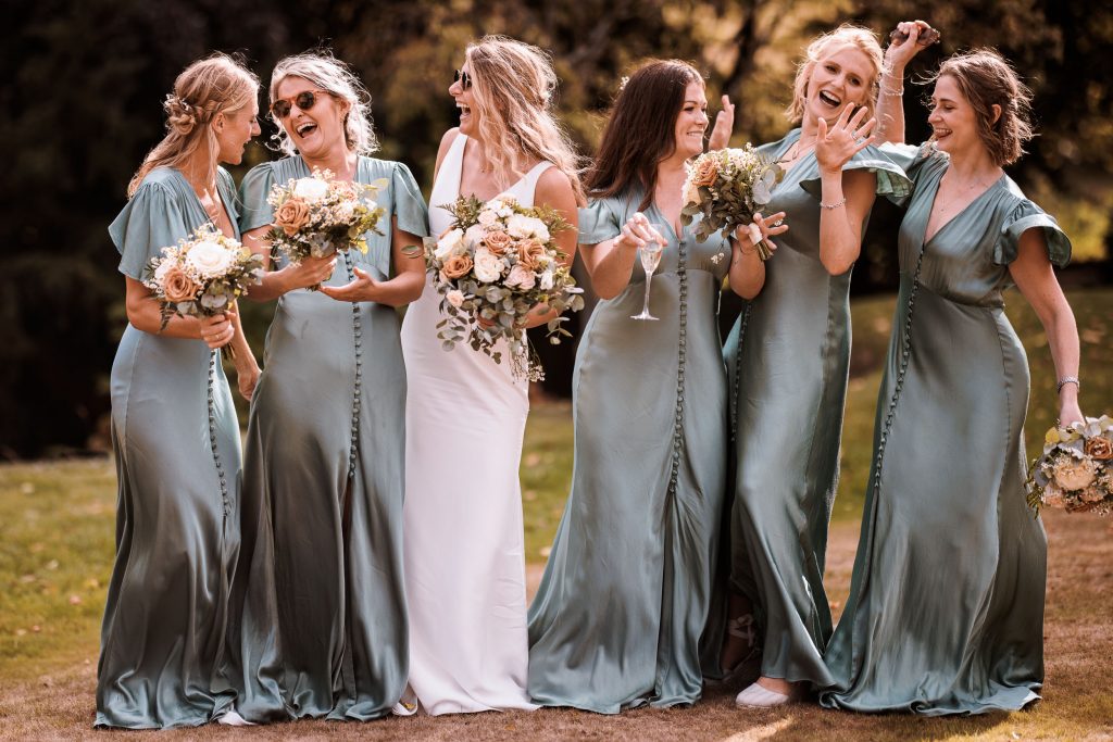 Sarah posses with her bridesmaids. Sarah wears a bespoke wedding dress and her bridesmaids are in satin long green dresses with button detailing down the centre front. 