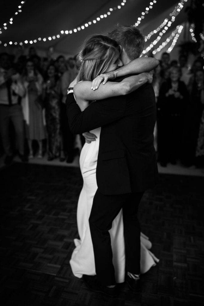 Black and white image. Sarah and George embrace as they have their first dance under the twinkly fairy lights inside the marquee.