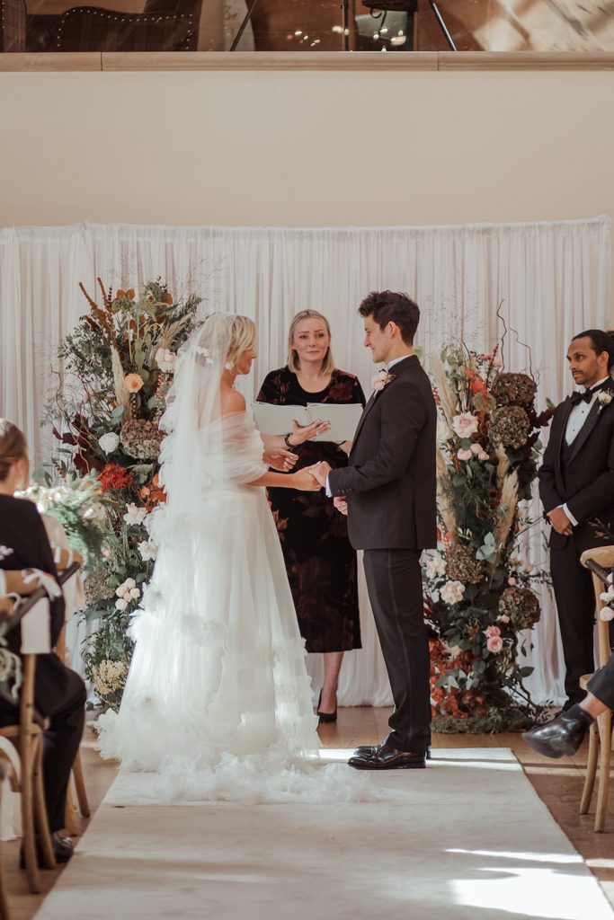 Laura and Edmund during their celebrant wedding ceremony. The top of the aisle is dressed with a white curtain and 2 large autumnal floral pillars.