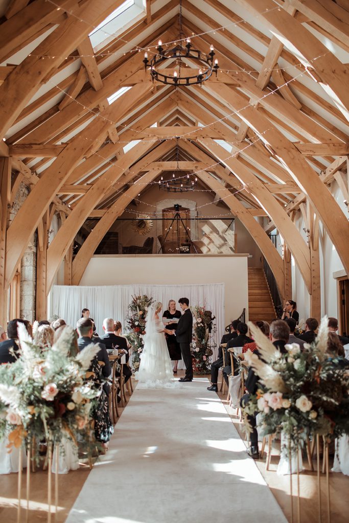 Laura and Edmund marry. Wide shot of the barn with guests seated during the ceremony. Exposed wooden beams, crossback chairs dressed with white chair sashes and beautiful autumnal florals complete the look.