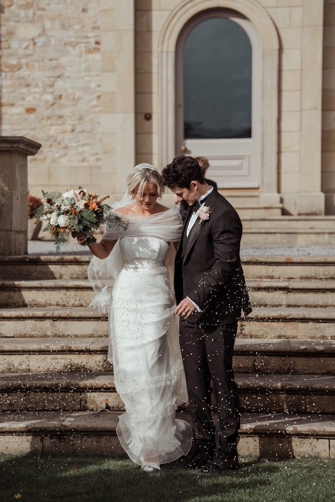 Laura and Edmund walk down the steps to the lawn for their drinks reception. Laura wears a beautiful Halfpenny London wedding dress and Edmund wears a black tux.