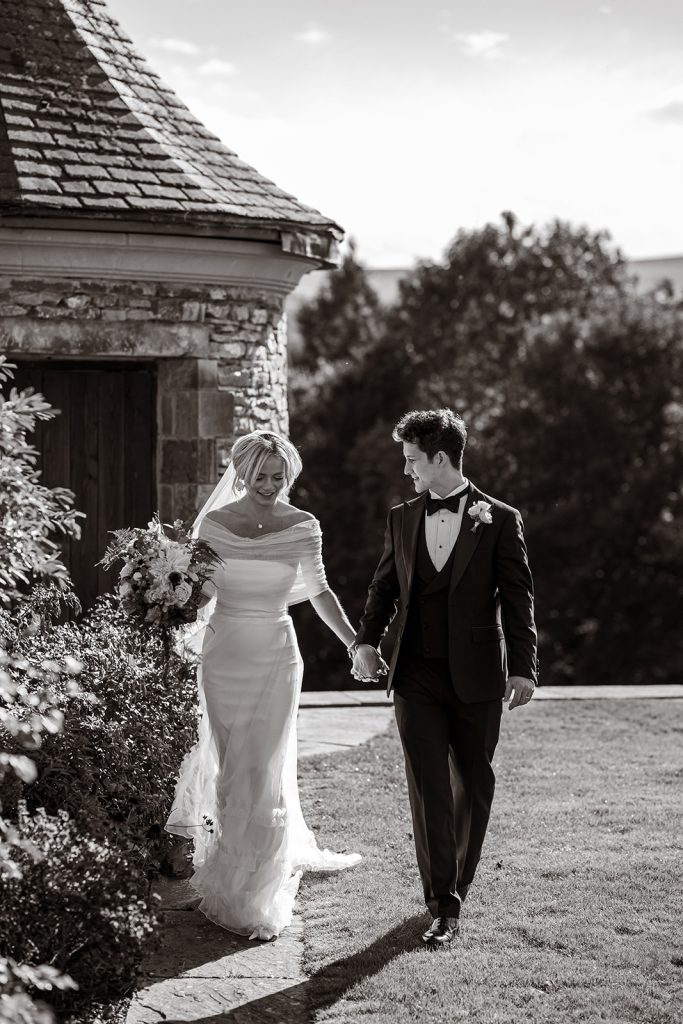 Black and white image. Laura and Edmund stroll across the lawn during their couple portraits. A stone corner house and trees make up the backdrop.