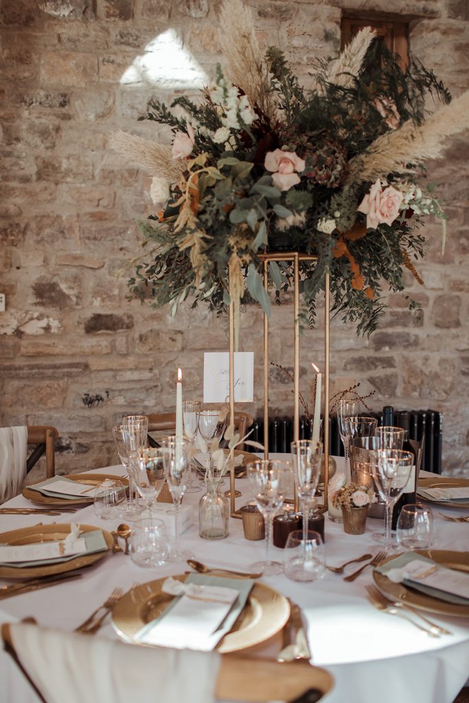 Close up of the wedding breakfast tables. Large autumnal florals with pampas grass sit on tall gold geometric stands. The table is decorated with gold charger plates, gold cutlery and gold rimmed glasses and finished with sage green napkins and hand made stationery.