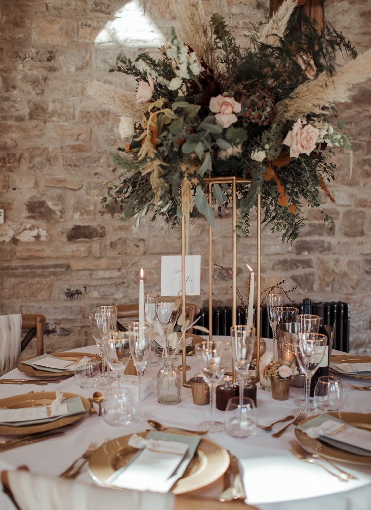 Wedding breakfast table dressed with a tall autumnal floral arrangement, gold charger plates, gold cutlery and elegant glassware.