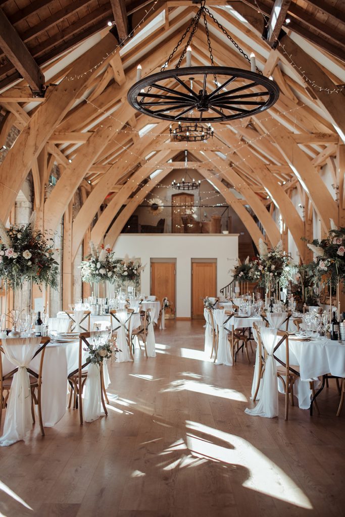 The barn at Tupgil Park is decorated ready for the wedding breakfast. The barn is filled with round tables dressed in white linen and large autumnal blooms. Cross back chairs and finished with white chair sashes.