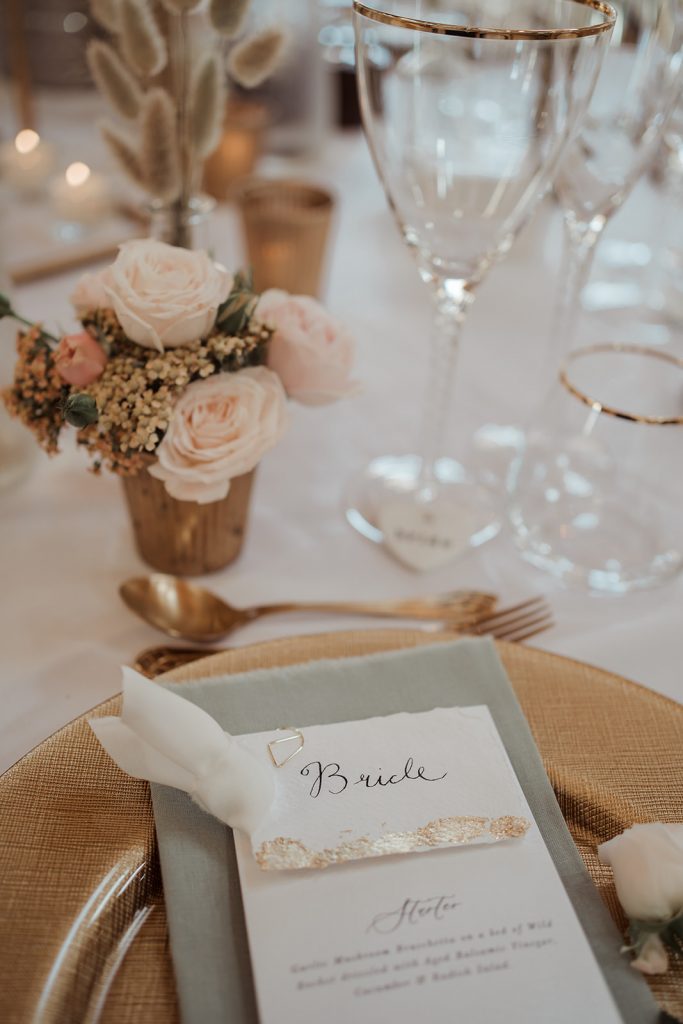 Close up shot of the table settings. Handmade stationery is finished with gold leaf and set upon a sage green napkin. Each setting has a gold charger plate, gold cutlery and gold rimmed glassware. Little poesy's of beautiful soft pink roses finish off the table.