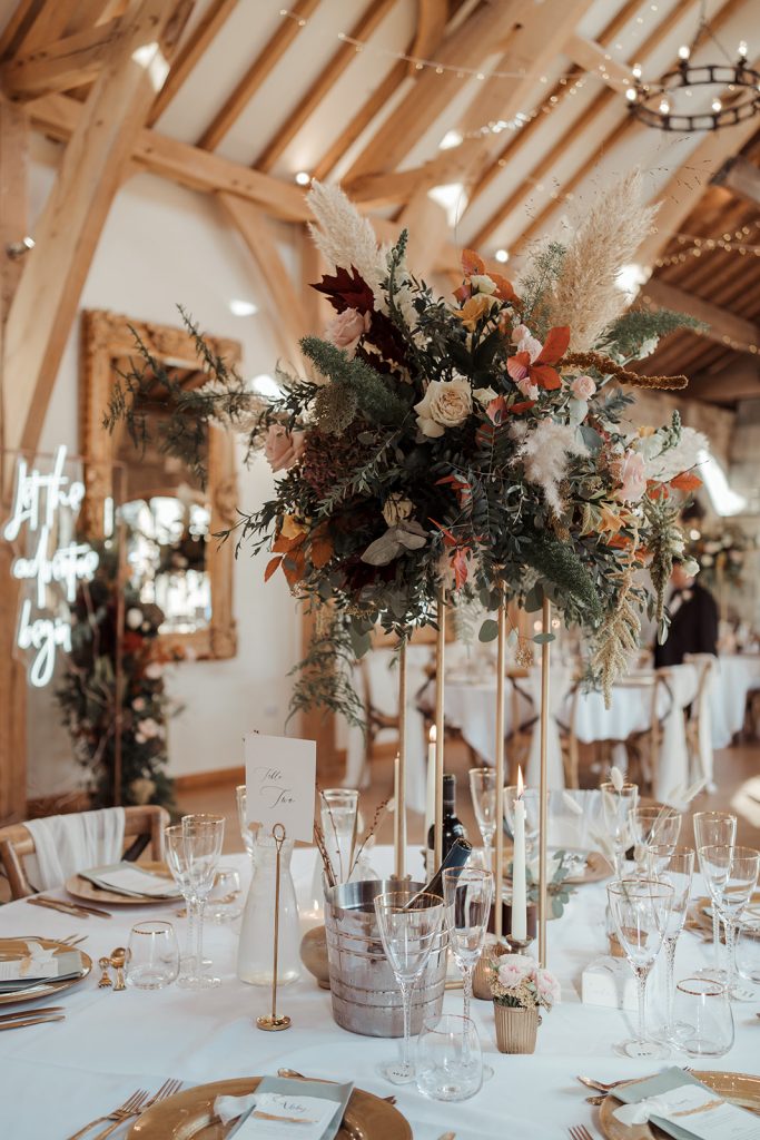Close up of the wedding breakfast tables. Large autumnal florals with pampas grass sit on tall gold geometric stands. The table is decorated with gold charger plates, gold cutlery and gold rimmed glasses and finished with sage green napkins and hand made stationery.