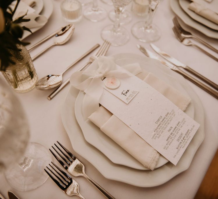 Wedding menu and name card created by Herve Jones Designs using seed paper. Stationery is placed upon a oatmeal napkin and scalloped edged plates. Silver cutlery and bud vases finish off the settings.