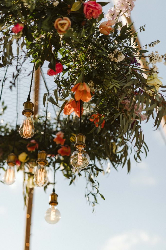 Large floral hoop consisting of bright pink, orange and yellow flowers with Eddison bulbs hangs from a naked tipi. Beautiful florals without the environmental impact.