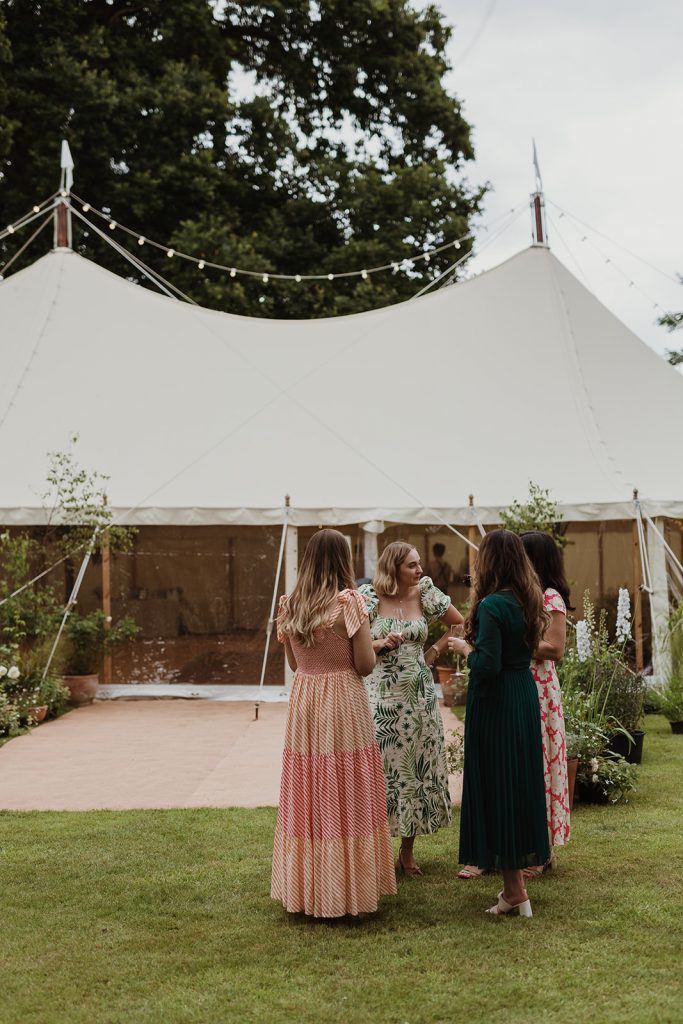 Marquee drinks reception held in a private garden. Guests stand in front of a beautiful sailcloth marquee chatting and drinking during the marquee drinks reception. Potted plants are used to decorate the entrance to the marquee.
