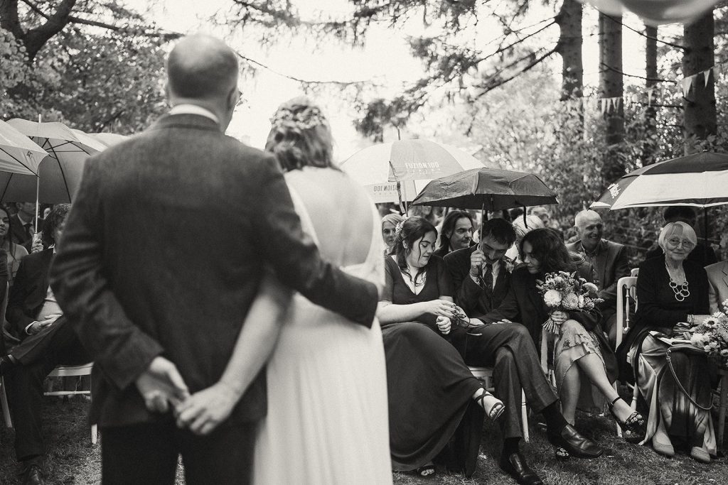 A black-and-white photo of an outdoor wedding. A couple stands with their backs to the camera facing seated guests. It appears to be raining, as many guests hold umbrellas as their wet weather plan. The scene feels intimate and timeless, set among trees with a calm, emotional atmosphere.
