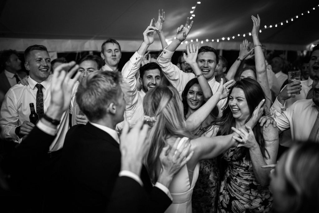 Black and white image of the bride and groom surrounded by their friends on the dancefloor. They have their arms in the air cheering and smiling.